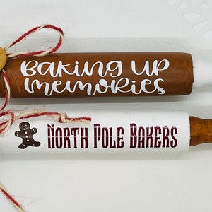May include: Two white wooden rolling pins with brown wooden handles. The top rolling pin has the text "baking up memories" in white lettering. The bottom rolling pin has the text "North Pole Bakers" in maroon lettering and a gingerbread man graphic.