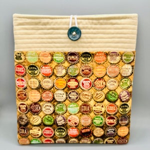 May include: A beige fabric pouch with a blue button closure. The pouch is lined with a fabric featuring a vintage pattern of colorful bottle caps with various soda brand names.