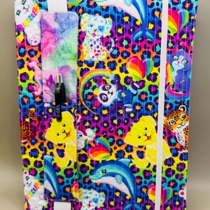 May include: A colorful notebook cover with a whimsical design featuring cartoon animals, rainbows, and hearts on a leopard print background. It has a pen holder and an elastic band closure. The cover is approximately 9 inches tall and 7 inches wide.