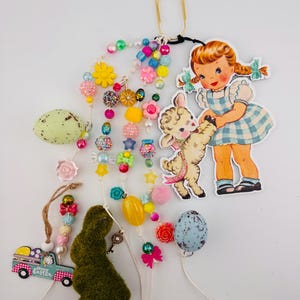 May include: A colorful Easter-themed garland with beads, flowers, candy, a lamb, a girl, and a bunny. The garland is attached to a gold paper clip. The garland also includes a green bunny, a blue speckled egg, and a yellow egg.