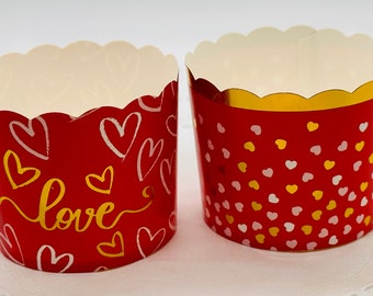 May include: Two red cupcake liners with gold accents. One liner has a white "love" inscription and white heart designs. The other liner has a scattered pattern of pink and gold hearts.