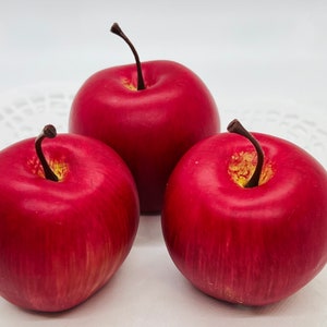 May include: Three vibrant red apples with dark brown stems and yellow accents. The apples are arranged closely together, showcasing their smooth, glossy surfaces. The image is set against a white background, highlighting the fruit's rich color and texture.