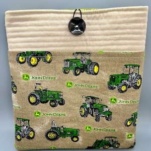 May include: A beige fabric tote bag with a black button closure. The bag is lined with a brown fabric featuring a repeating pattern of green John Deere tractors.