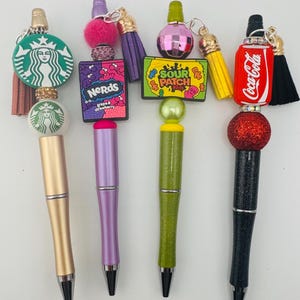 May include: Four decorative pens with different themed charms. The first pen is gold with a Starbucks logo charm. The second pen is purple with a Nerds candy charm. The third pen is green with a Sour Patch Kids charm. The fourth pen is black with a Coca-Cola charm.