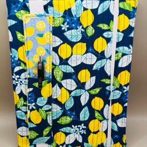 May include: A fabric journal cover with a vibrant lemon pattern on a navy blue background. The cover features a pocket with a lemon slice design and a black pen. A white elastic band secures the journal.