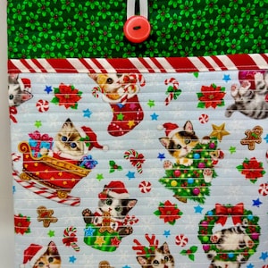 May include: A quilted fabric pouch featuring a light blue background with a Christmas-themed print of kittens, candy canes, poinsettias, and gingerbread men. The top flap is green with holly leaves and secures with a red button and elastic loop.