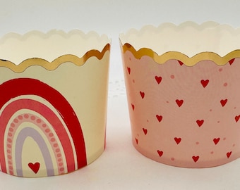 May include: Two cupcake liners, one with a rainbow design and the other with a pink background and red hearts. Both liners have a gold scalloped edge.