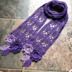 May include: A purple crocheted scarf with a gradient of color from light to dark. The scarf has a delicate lace pattern and features three crocheted flowers.