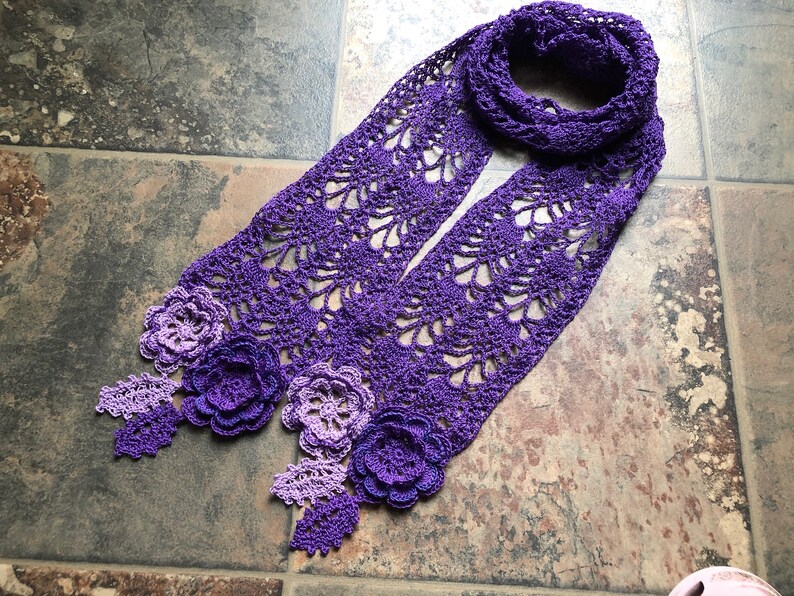 May include: A purple crocheted scarf with a gradient of color from light to dark. The scarf has a delicate lace pattern and features three crocheted flowers.