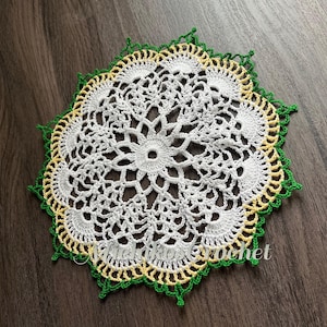 May include: White crochet doily with a green and yellow border. The doily has a delicate floral design.