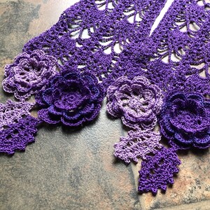 May include: A purple crocheted scarf with a gradient of color from light to dark. The scarf has a delicate lace pattern and is adorned with three crocheted flowers.