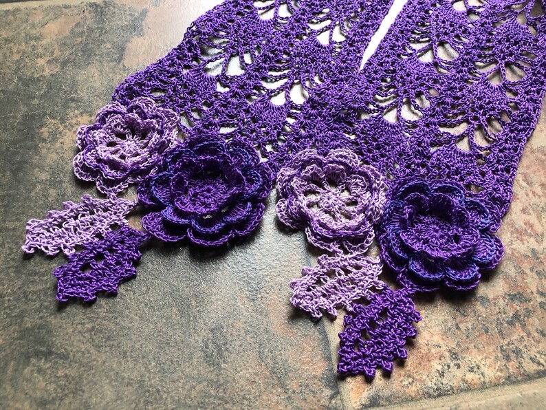 May include: A purple crocheted scarf with a gradient of color from light to dark. The scarf has a delicate lace pattern and is adorned with three crocheted flowers.