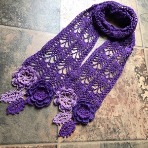 May include: A purple and lavender crocheted scarf with a delicate floral design. The scarf is made with a lacy stitch and features several crocheted flowers.