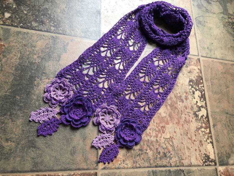 May include: A purple and lavender crocheted scarf with a delicate floral design. The scarf is made with a lacy stitch and features several crocheted flowers.