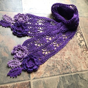 May include: A purple crocheted scarf with a delicate lace pattern and flower embellishments. The scarf is made with a gradient of purple yarn, starting with a lighter shade and transitioning to a darker shade.