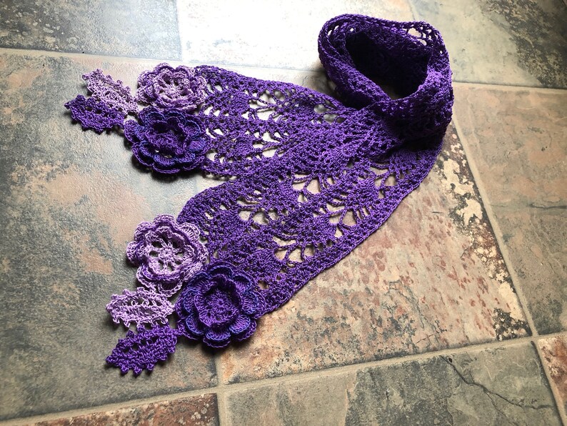 May include: A purple crocheted scarf with a delicate lace pattern and flower embellishments. The scarf is made with a gradient of purple yarn, starting with a lighter shade and transitioning to a darker shade.