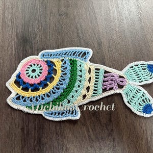 May include: A colorful, crocheted fish in shades of blue, pink, yellow, green, and purple. The fish has a detailed, openwork design with a white border. The text "michiko crochet" is visible on the fish.