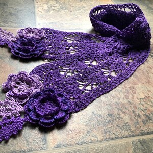 May include: A purple and lavender crocheted scarf with flower embellishments. The scarf is made with a delicate openwork pattern and features three dimensional flowers.