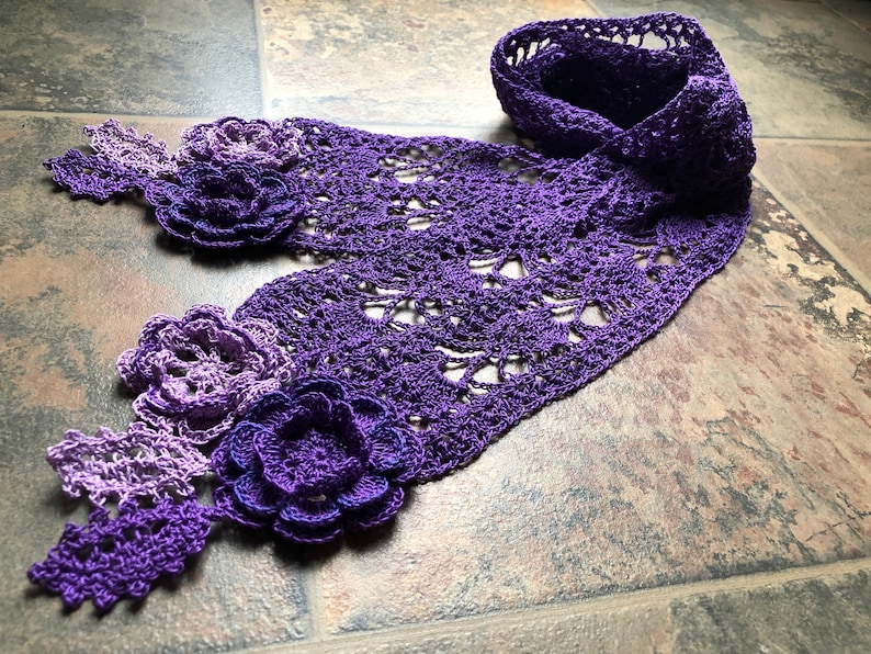 May include: A purple and lavender crocheted scarf with flower embellishments. The scarf is made with a delicate openwork pattern and features three dimensional flowers.