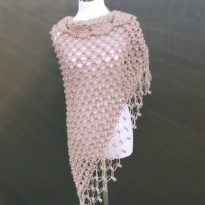 May include: A beige, open-weave shawl with a scalloped neckline and fringed edges. The shawl is displayed on a white mannequin against a muted green background. The knitted design creates a delicate, airy appearance.