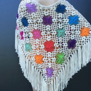 May include: A white crochet poncho with colorful flower accents and fringe trim. The poncho is made with a delicate openwork stitch and features a variety of bright colors, including red, orange, yellow, green, blue, purple, and pink.