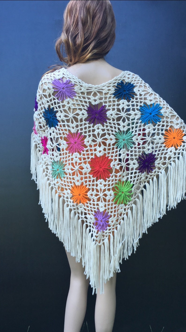 May include: A white crochet poncho with colorful flower accents and fringe trim. The poncho is made with a delicate openwork stitch and features a variety of bright colors, including red, orange, yellow, green, blue, purple, and pink.
