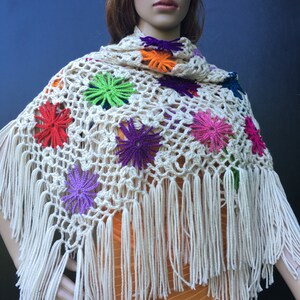 May include: A white crocheted shawl with colorful flower accents and fringe. The shawl is draped over a mannequin.
