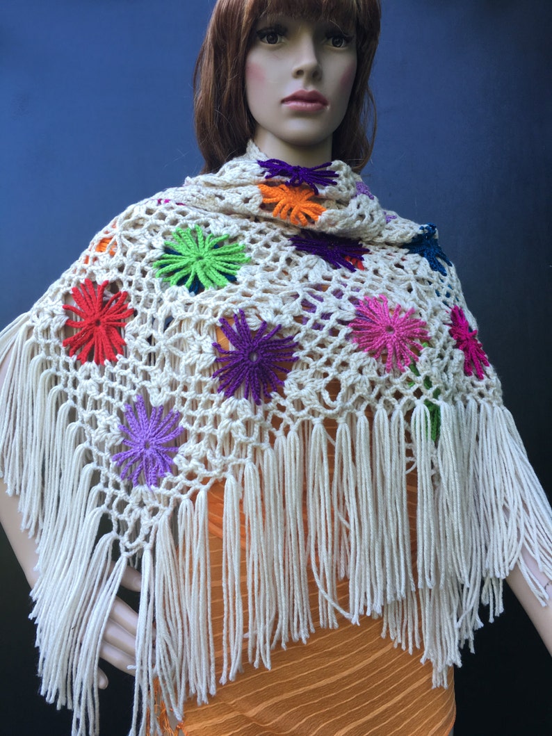 May include: A white crocheted shawl with colorful flower accents and fringe. The shawl is draped over a mannequin.
