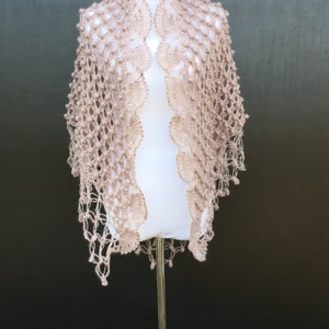 May include: A light brown, open-weave crochet shawl displayed on a white mannequin. The shawl features a delicate, lacy pattern with scalloped edges and a fringe. The mannequin is on a metal stand against a dark background.