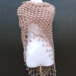 May include: A light brown, open-weave shawl with a scalloped neckline and fringe detail. The shawl is displayed on a white mannequin against a dark background. The crochet design features a repeating pattern of small, raised circles.