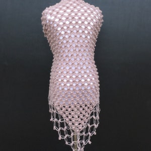 May include: A light brown, open-weave crochet top displayed on a mannequin. The top has a net-like pattern with a high neckline and a fringed hem. The garment is made of a soft, textured yarn, and the overall style is bohemian.