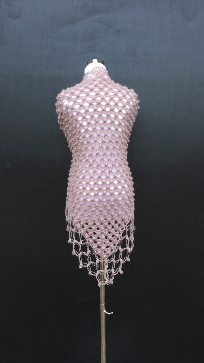 May include: A light brown, open-weave crochet top displayed on a mannequin. The top has a net-like pattern with a high neckline and a fringed hem. The garment is made of a soft, textured yarn, and the overall style is bohemian.