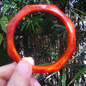 Op de afbeelding: Een ronde armband met een gefacetteerde, achthoekige buitenrand. De armband is gemaakt van een doorschijnend, gevlekt materiaal in tinten diep oranje, amber en roodbruin, met een gladde, gepolijste afwerking over het gehele oppervlak.