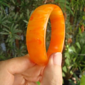 Puede incluir: Una pulsera tipo brazalete lisa y pulida que presenta un tono naranja vibrante y translúcido con patrones de remolinos de tonos más claros y oscuros en todo el material, creando un efecto jaspeado en la superficie redondeada de la pieza circular.