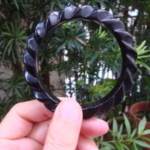 May include: A circular black bangle bracelet featuring a continuous twisted or rope-like carved pattern along its entire outer surface. The bangle has a smooth, polished finish and a uniform thickness, creating a distinct ridged texture.