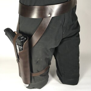 May include: A brown leather thigh holster with a black handgun. The holster is attached to a brown leather belt with straps that go around the legs.