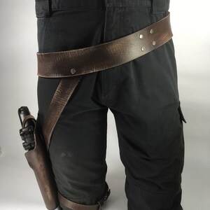 May include: A brown leather belt with metal rivets is worn around a pair of black cargo trousers. A brown leather holster with a silver gun is attached to the belt and worn on the left thigh.