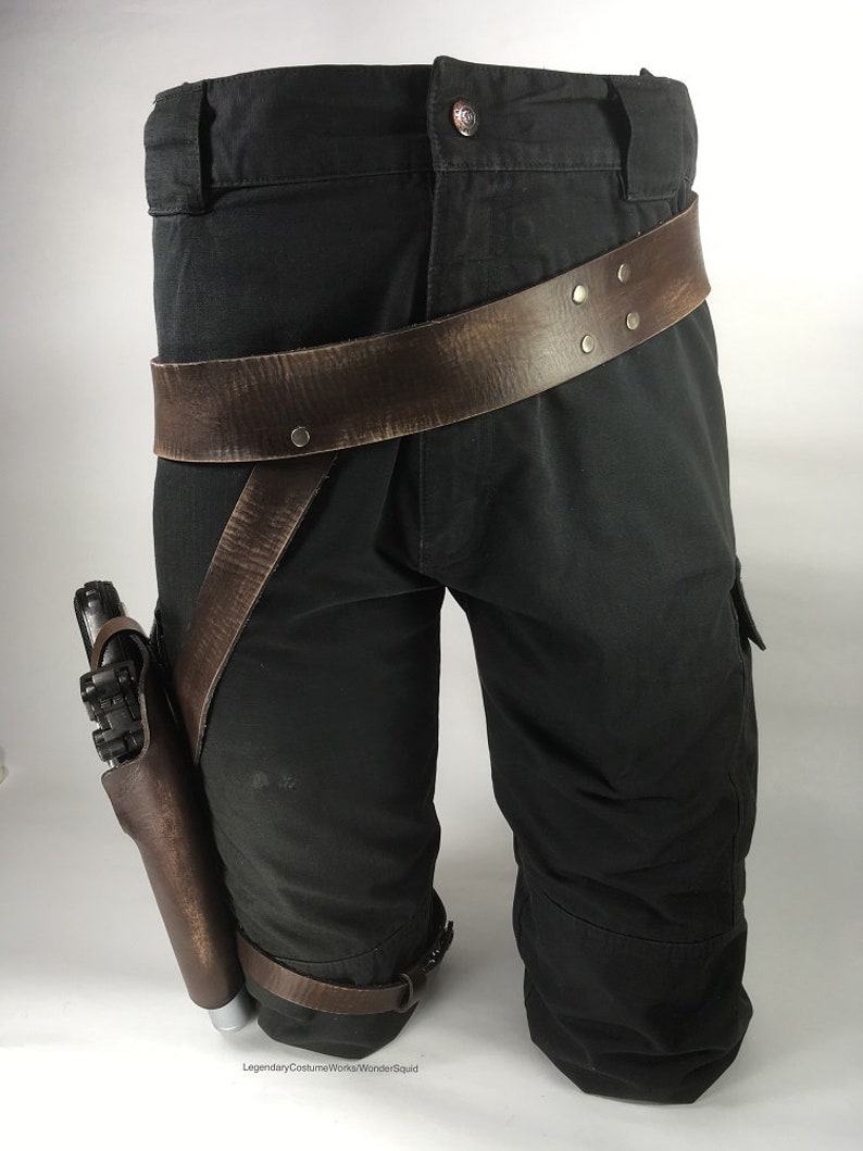 May include: A brown leather belt with metal rivets is worn around a pair of black cargo trousers. A brown leather holster with a silver gun is attached to the belt and worn on the left thigh.