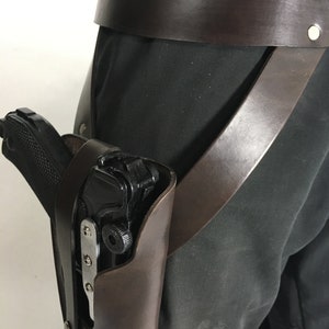 May include: A brown leather holster with a black gun inside. The holster is attached to a brown leather belt with a metal buckle.