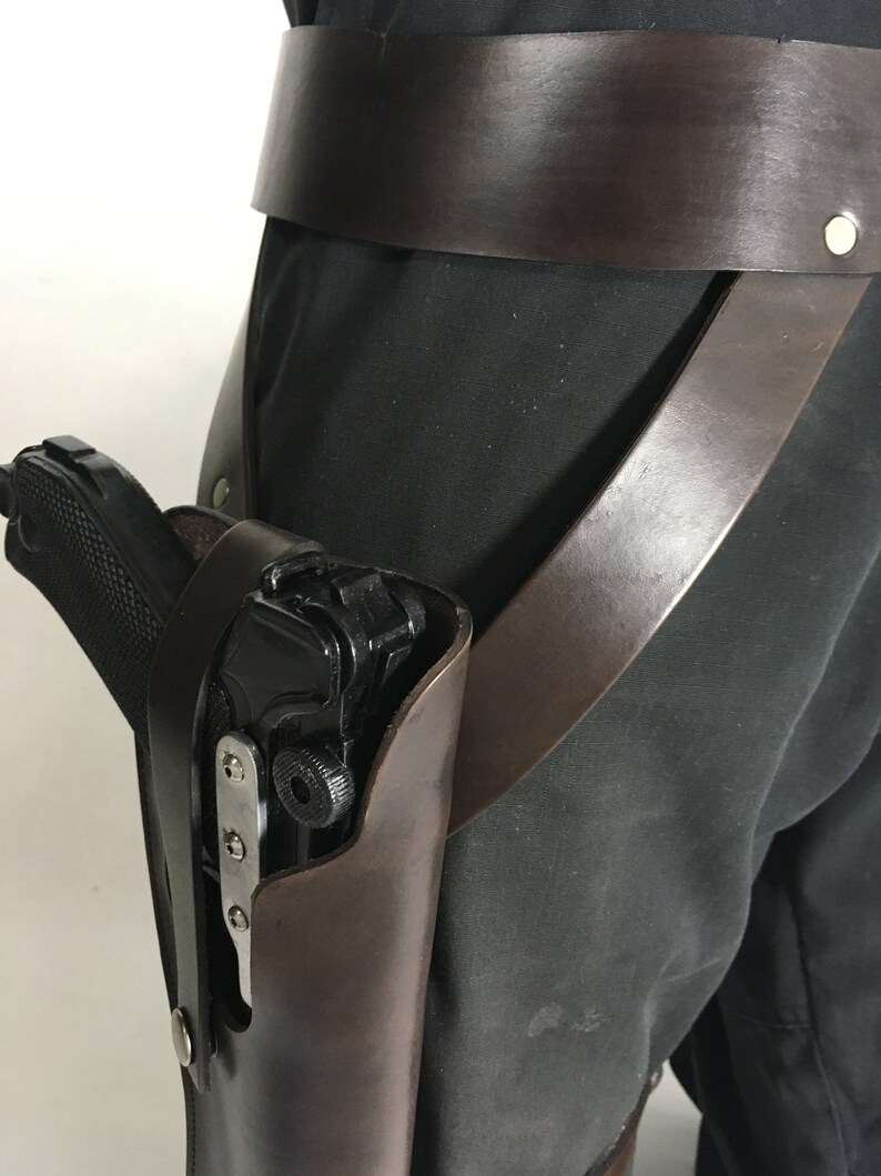 May include: A brown leather holster with a black gun inside. The holster is attached to a brown leather belt with a metal buckle.