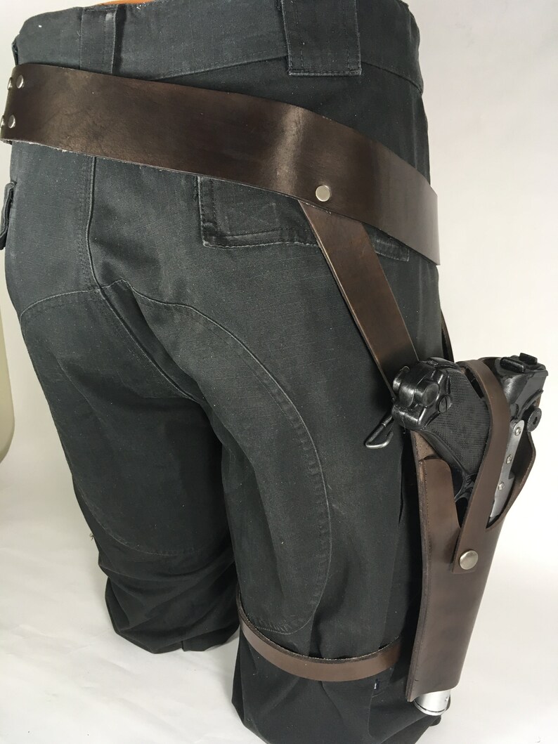May include: A brown leather holster with a gun attached to it. The holster is worn on the leg and secured with a brown leather strap. The holster is designed to be worn on the right leg.