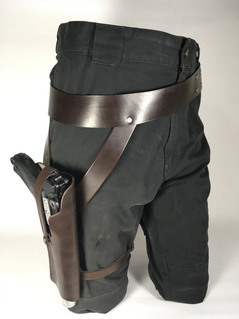 May include: A brown leather thigh holster with a black handgun. The holster is attached to a brown leather belt with straps that go around the legs.