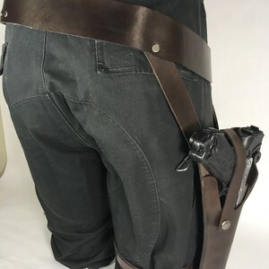 May include: A brown leather holster with a black gun attached to a brown leather belt. The holster is worn on the right leg and is secured with a strap that goes around the thigh.