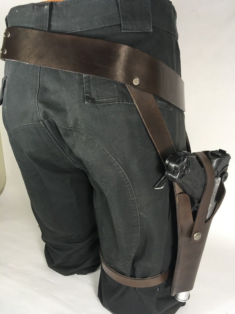 May include: A brown leather holster with a black gun attached to a brown leather belt. The holster is worn on the right leg and is secured with a strap that goes around the thigh.