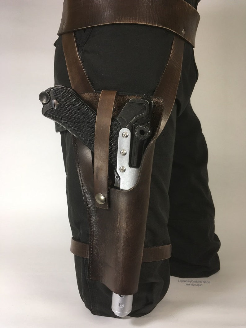 May include: A brown leather holster with straps and buckles, worn on a person's leg, holding a black and silver pistol.