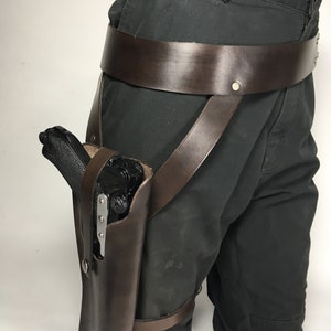 May include: A brown leather thigh holster with a black gun inside. The holster is attached to a brown leather belt with straps that go around the leg.