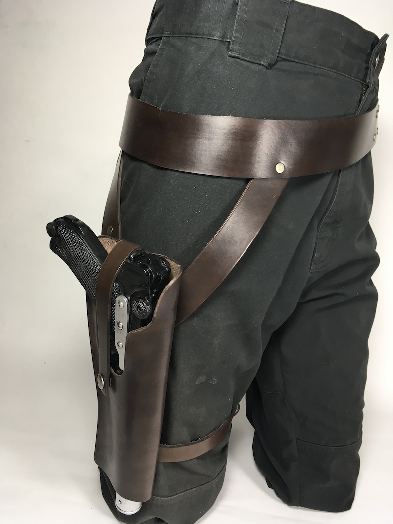 May include: A brown leather thigh holster with a black gun inside. The holster is attached to a brown leather belt with straps that go around the leg.
