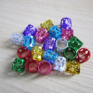 Op de afbeelding: Een verzameling kleurrijke metalen haar kralen in verschillende tinten, waaronder paars, blauw, groen, rood, zilver en goud. Elke kraal heeft een cilindrische vorm met decoratieve uitgesneden patronen. De kralen zijn verspreid op een lichtgekleurd houten oppervlak.