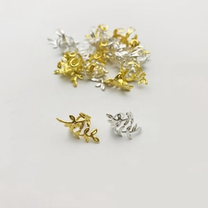 May include: A collection of small, decorative metal charms in gold and silver tones. The charms feature a floral and leaf design. The charms are scattered on a white surface.