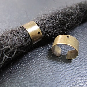 Op de afbeelding: Twee goudkleurige metalen dreadlock kralen met een klein gaatje in elk. De kralen zitten op een zwarte dreadlock.